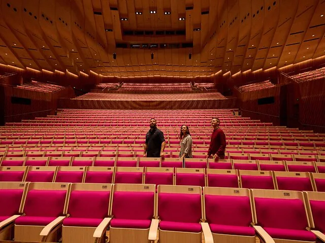 Drie mensen staan tussen de vele lege rode stoelen in een zaal.